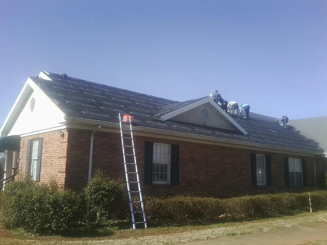Crew tearing shingles off a brick ranch roof with a ladder set — Metal Roof Repair work by our crew in Fort Jennings