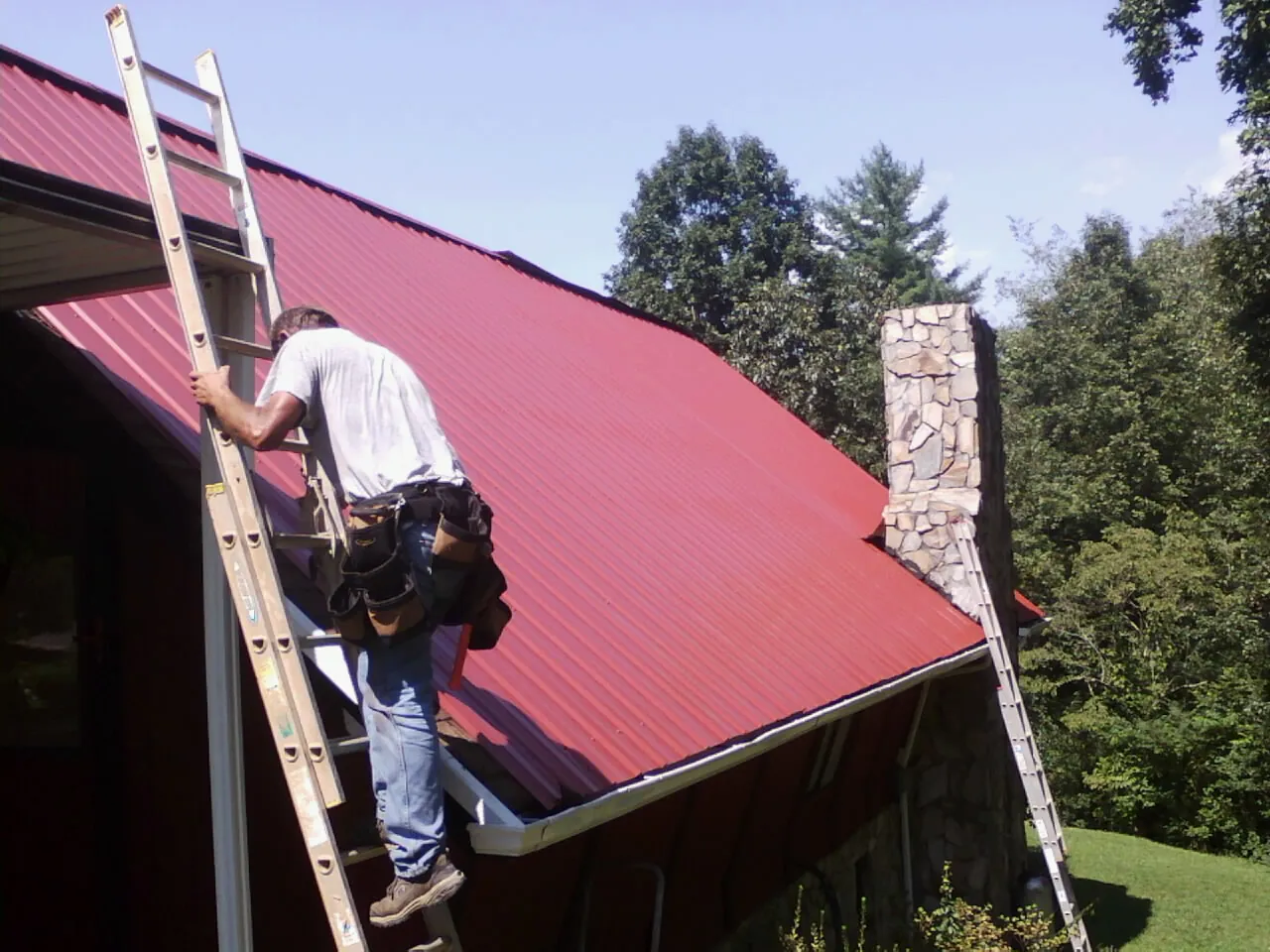 Roofer on a ladder beside a red metal roof and stone chimney — Soffit & Fascia Repair by Roofing Brothers in Fort Jennings